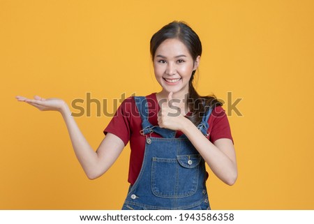 Portrait close up half-length shot of beautiful Asian woman in a denim dungarees. She point her hand to the other hand for add some product or advertising on her hand isolated on yellow background.