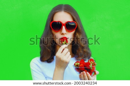 Portrait close up of happy smiling woman with handful of fresh strawberries wearing a red heart shaped sunglasses on green background
