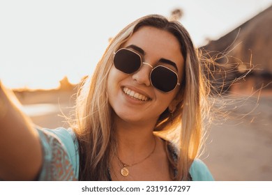 Portrait close up of one young beautiful attractive blonde young girl holding camera and taking selfie picture at the beach enjoying outdoors free time with sunset in the background. - Powered by Shutterstock