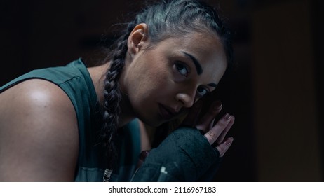 Portrait Close Up Shot Of A Badass Confident Athletic Female Wearing Gloves With Braids Looking Directly At The Camera With A Black Background. Confident Looking Kick Boxer Taking A Break .