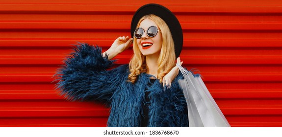 Portrait close up of stylish blonde woman with two shopping bags wearing a blue faux fur coat, black round hat over red wall background - Powered by Shutterstock
