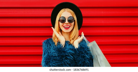 Portrait close up of surprised young woman with shopping bags wearing blue faux fur coat, black round hat over red wall background