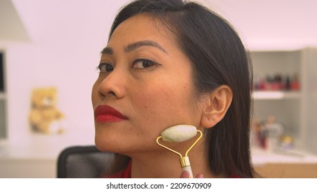 PORTRAIT: Close Up View Of Young Lady Self Massaging Her Face With Jade Scraper. Beautiful Asian Woman Using Gua Sha Scraper For Rejuvenating Anti-aging Massage And Facial Muscles Relief Treatment.