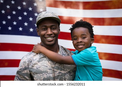 Portrait Close Up Of A Young Adult African American Male Soldier Carrying His Young Son, Both Looking To Camera And Smiling In Front Of A US Flag