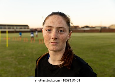 Portrait close up of a young adult Caucasian female rugby player standing on a rugby pitch looking to camera, with her teammates training in the background - Powered by Shutterstock