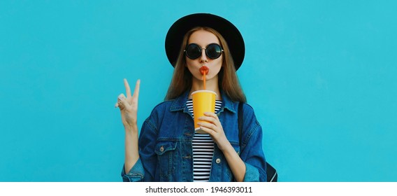 Portrait close up of young woman drinking a juice wearing a black round hat, denim jacket on a blue background