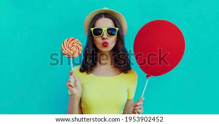 Portrait close up of young woman with lollipop and red balloon blowing a red lips wearing a summer straw hat on a blue background
