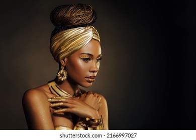 Portrait Closeup Beauty Fantasy African Woman, Face In Gold Paint. Golden Shiny Black Skin. Fashion Model Girl Mixed Race. Glamorous Arab Turban, Jewellery Accessories. Professional Metallic Makeup.