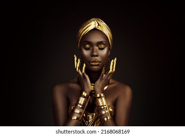 Portrait closeup Beauty fantasy african woman face in gold paint. Golden shiny skin. Fashion model girl goddess hand fingers posing. Arab turban head, jewellery bracelets. Professional metallic makeup