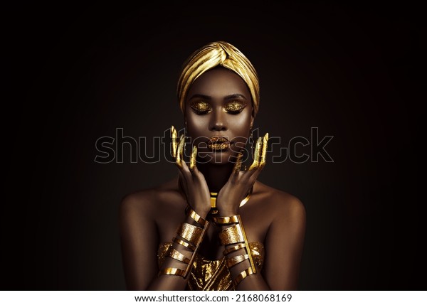 Portrait closeup Beauty fantasy african woman face
in gold paint. Golden shiny skin. Fashion model girl goddess hand
fingers posing. Arab turban head, jewellery bracelets. Professional
metallic makeup