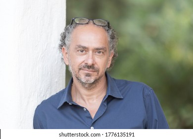 Portrait Closeup Of A Middle Aged Italian Man With Beard And His Graying Curly Hair Blue Shirt And Jeans Leaning Against A White Wall