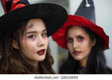 Portrait Closeup Shot Of Asian Young Scary Creepy Female Model In Tall Hat Horror Witch Costume Wearing Scar Makeup With Friend In Blurred Background In Traditional Halloween Trick Or Treat Party.