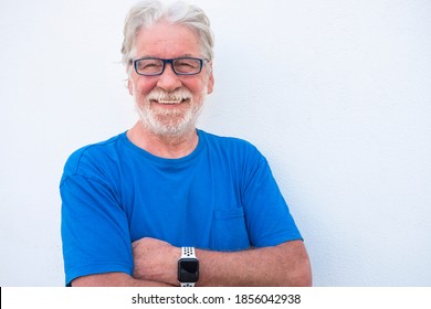 Portrait Close-up Of A Smiling Senior Man With White Beard On White Background Wearing Eyeglasses And Tshirt In Blue Color. Positive Retiree Person With Crossed Arms Looking At Camera