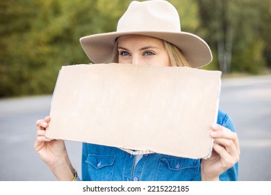 Portrait Closeup Of Woman Hopefully Waiting Passing Car In Forest Holding Empty Cardboard Poster On Road. Lady In Hat Escape From City By Auto Stop To Go Anywhere. Travelling, Hitchhiking, Vacations.