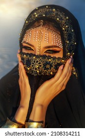 portrait close-up, young beautiful oriental woman. Beauty girl face hidden by golden veil head covered black scarf. Luxurious evening arabic style make-up, smoky green eyes. golden shadows