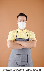 Portrait Of Coffeeshop Barista In Apron And Medical Mask Crossing Arms And Looking At Camera