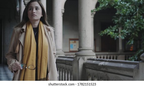 Portrait Of College Student Walking In Colonnade Hallway. Young Lady In Coat Looking Away In University Building. Successful Businesswoman Putting On Eyeglasses Outside In Slow Motion