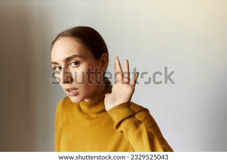 Portrait of concentrated anxious female eavesdropping holding head next to ear, listening carefully to strange sounds, feeling scared, standing against gray studio background with opened mouth