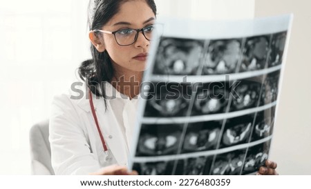 Portrait of concentrated indian woman doctor therapist with glasses thinking about diagnosis while looking at results patient MRI or CT scan procedure at light hospital office