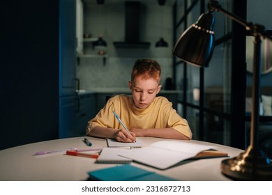 Portrait of concentrated pupil boy studying at home doing homework sitting at table under light of lamp at evening. Front view of schoolboy writing in exercise book at night in dark with lighting lamp - Powered by Shutterstock