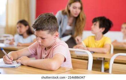 Portrait Of Concentrated Tween Boy Studying In Classroom, Listening To Schoolteacher And Writing In Notebook