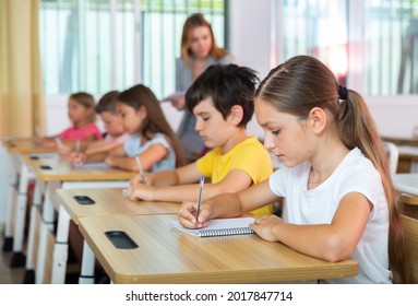 Portrait Of Concentrated Tween Girl Studying In Classroom, Listening To Schoolteacher And Writing In Notebook