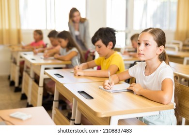 Portrait Of Concentrated Tween Girl Studying In Classroom, Listening To Schoolteacher And Writing In Notebook