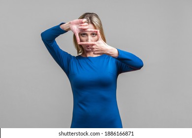 Portrait Of Concentrated Woman In Elegant Tight Blue Dress Looking At Camera Through Hand Frame, Capturing Photo Or Focusing Eyesight On Target. Indoor Studio Shot Isolated On Gray Background