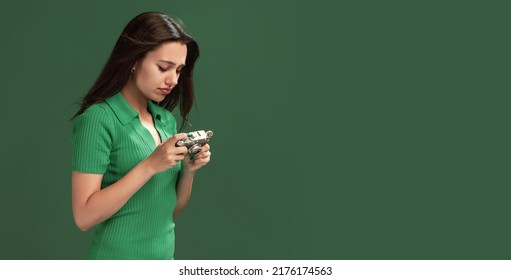 Portrait Of Concentrated Young Girl Looking At Photos In Retro Camera Isolated Over Green Studio Background. Memories. Concept Of Emotions, Facial Expression, Lifestyle, Fashion, Youth Culture