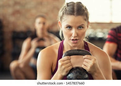 Portrait Of Concentrated Young Woman Working Out With Kettlebell At Gym. Closeup Face Of Determined Girl Doing Squat Session And Training Biceps. Fitness Class Lifting Heavy Weights While Squatting.