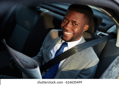 Portrait of confident African - American businessman riding in backseat of car  looking at camera out lit by sunlight, smiling cheerfully while reading newspaper - Shutterstock ID 614635499