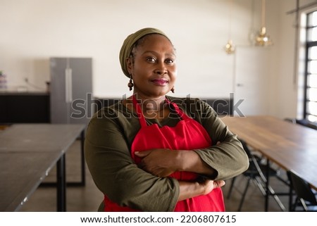 Portrait of a confident African American woman wearing a red apron at a cookery class, standing in a restauarant with her arms crossed, looking to camera and smiling