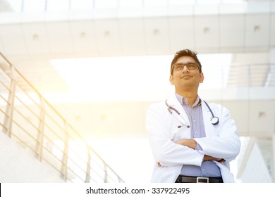 Portrait Of Confident Asian Indian Medical Doctor Standing Outside Hospital Building, Beautiful Golden Sunlight At Background.