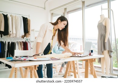 Portrait of a confident Asian tailor woman working on clothes in tailoring atelier. A young attractive female fashion designer smile and look at the camera with a dress-on mannequin in the background. - Powered by Shutterstock