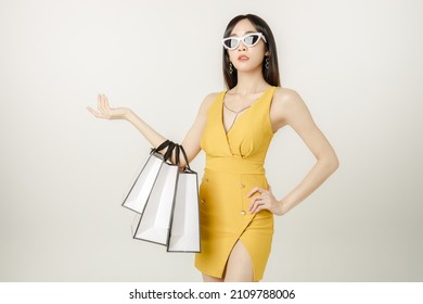 Portrait of Confident Asian woman in a sultry yellow dress wearing sunglasses and carrying a packages bag is having fun purchases after shopping isolated on white background.