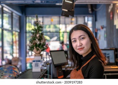 Portrait of confident Asian young adult owner gen z young woman at a counter bar in coffee shop cafe. She is wearing an apron looking at the camera and smiling. Hospitality and welcome concept.
