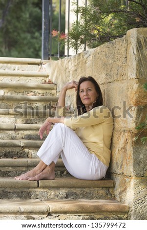 Portrait confident attractive mature woman sitting relaxed and happy smiling outdoor on limestone steps in park, bare feet and leisure, blurred background and copy space.