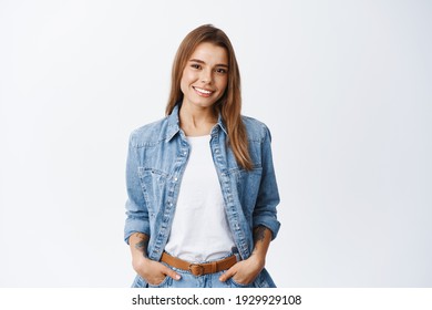 Portrait Of Confident Beautiful Woman With Short Hair, Wearing Casual Clothes, Standing In Relaxed Pose With Hands In Pockets, Smiling With White Teeth At Camera, Studio Background