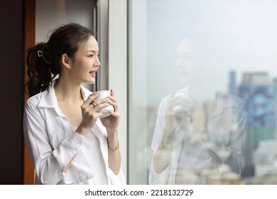 Portrait Of Confident And Beauty Young Asian Businesswoman With Coffee Cup Standing And Looking Away In Modern Hotel With City Blurred Background. Success Business And Lifestyle Concept