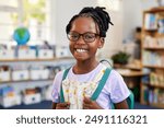 Portrait of a confident black girl wearing spectacles while holding backpack at library. Cheerful african american schoolgirl wearing eyeglasses standing in classroom while looking at camera. 