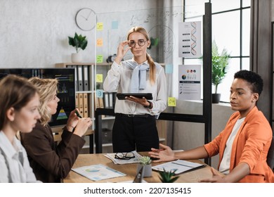 Portrait Of Confident Blond Woman In White Shirt And Eyewear Posing At Modern Office. Three Diverse Female Coworkers Talking Sitting Near Their Leader At Office.