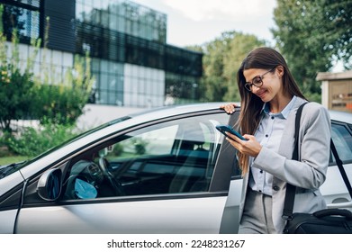 Portrait of a confident business woman wearing elegant suit and typing message on a smartphone while getting into the car. Businesswoman using mobile phone while standing next to open car door.