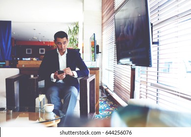 Portrait Of A Confident Businessman Using Smart Phone While Watching Something On TV During Recreation Time, Intelligent Man In Luxury Suit Holding His Cell Telephone While Sitting In Coffee Shop 