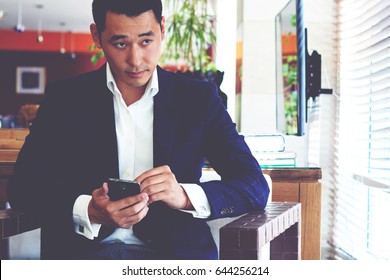 Portrait Of A Confident Businessman Using Smart Phone While Watching Something On TV During Recreation Time, Intelligent Man In Luxury Suit Holding His Cell Telephone While Sitting In Coffee Shop 