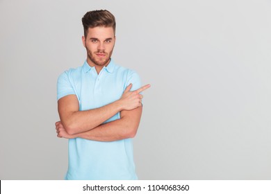 portrait of confident casual man wearing a light blue polo t-shirt pointing to side while standing on light grey background