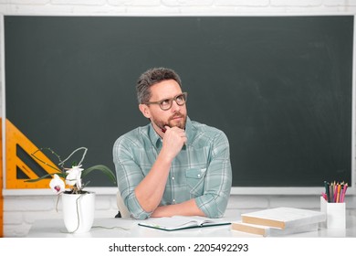 Portrait Of Confident Caucasian Male Teacher In Classroom. Young Male Thinking Teacher With Chalk Near Blackboard In Classroom.