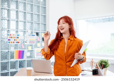 A portrait of a confident, cheerful Asian female graphic designer with red curly hair is standing in her studio with a graphic drawing tablet in her hand. - Powered by Shutterstock