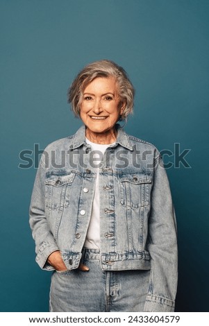 Portrait of a confident and cheerful mature woman with grey hair standing in a studio. She is wearing a fashionable denim outfit and a casual blue background complements her youthful and happy smile.