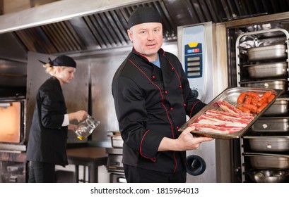 Portrait of confident chef working in restaurant kitchen, holding baking tray with raw meat products