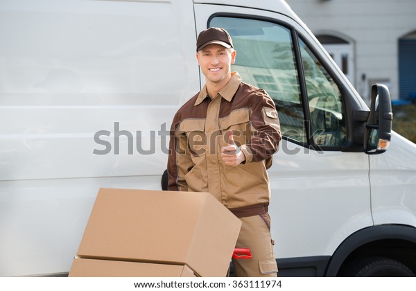 Portrait of confident delivery man pushing parcels on\
handtruck against van